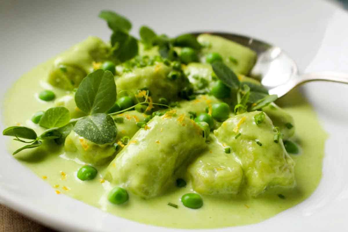 Close-up of spring pea gnocchi in a vibrant green sauce with scattered fresh peas and clover-shaped microgreens garnish.