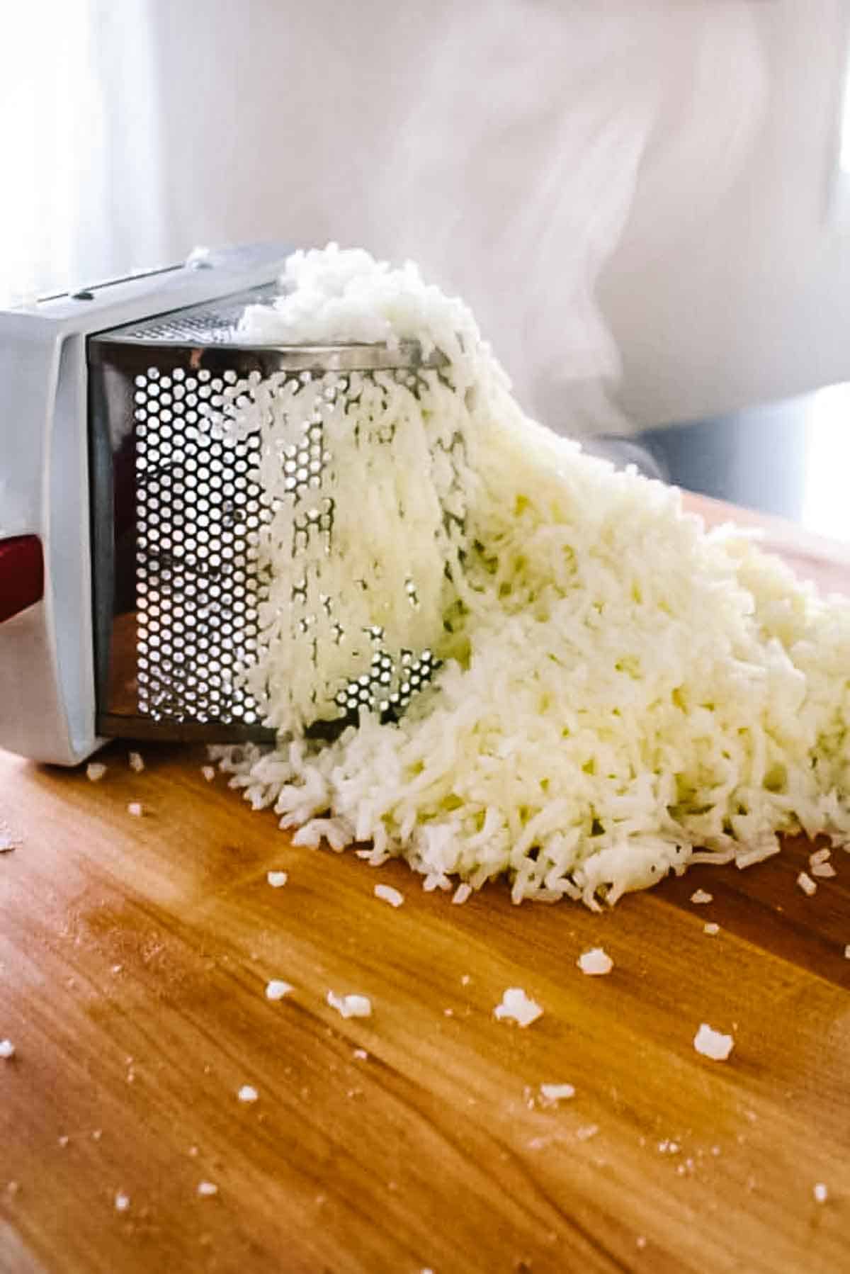 Box grater with freshly grated white potato for gnocchi dough on a wooden cutting board.