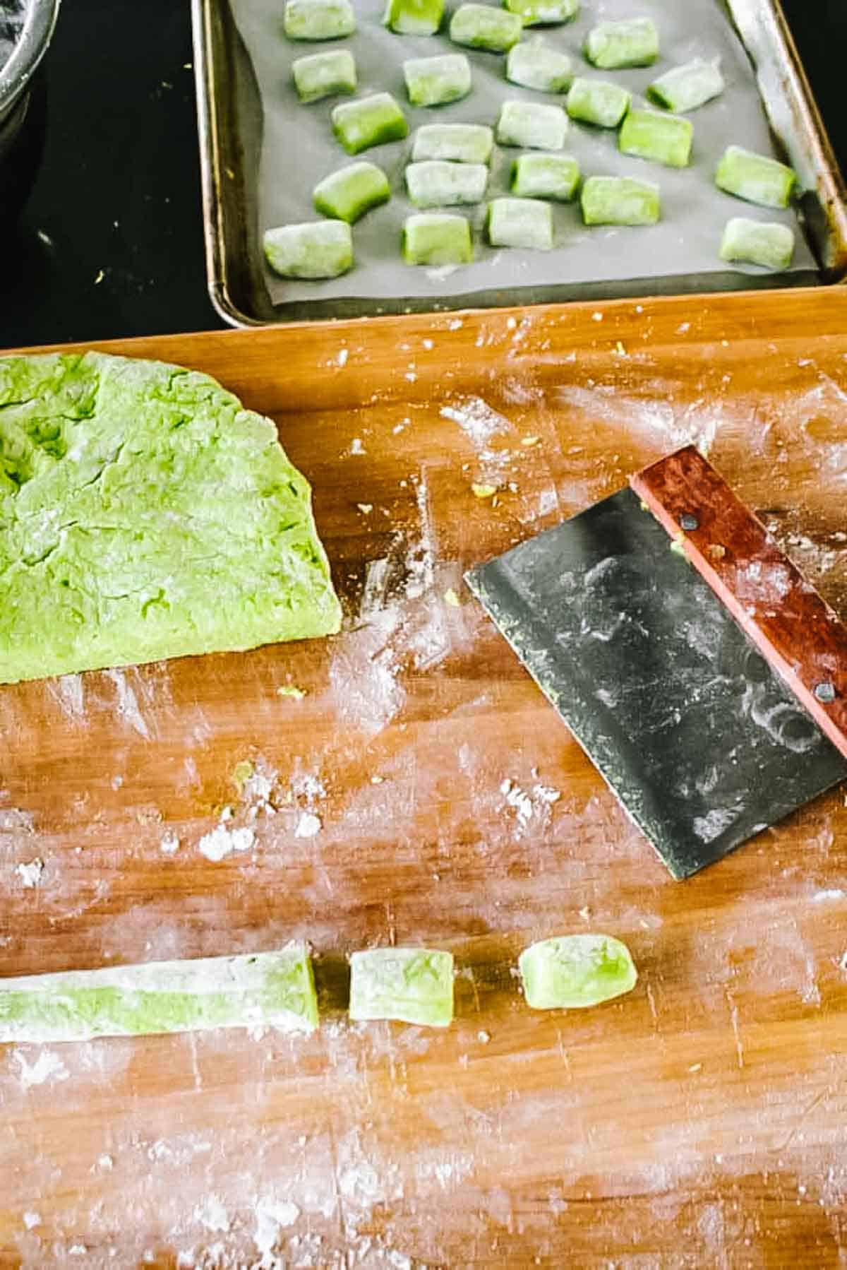 Green pea gnocchi dough being cut into small pillows on a floured wooden board with a bench scraper and formed pieces on a parchment-lined baking sheet.