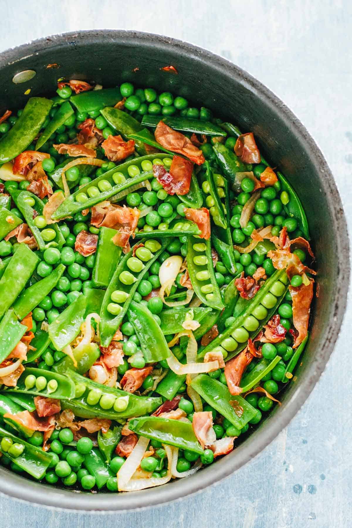 Overhead view of a dark pan filled with bright green peas and snow peas mixed with crispy prosciutto pieces and sliced onions.