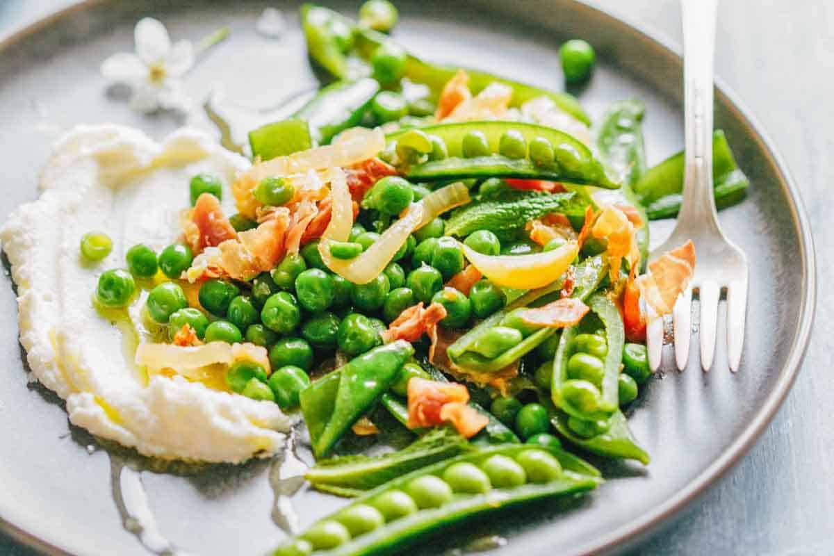 A serving of fresh peas and snow peas with crispy prosciutto and sautéed onions on a round plate with a dollop of creamy ricotta cheese and a silver fork.