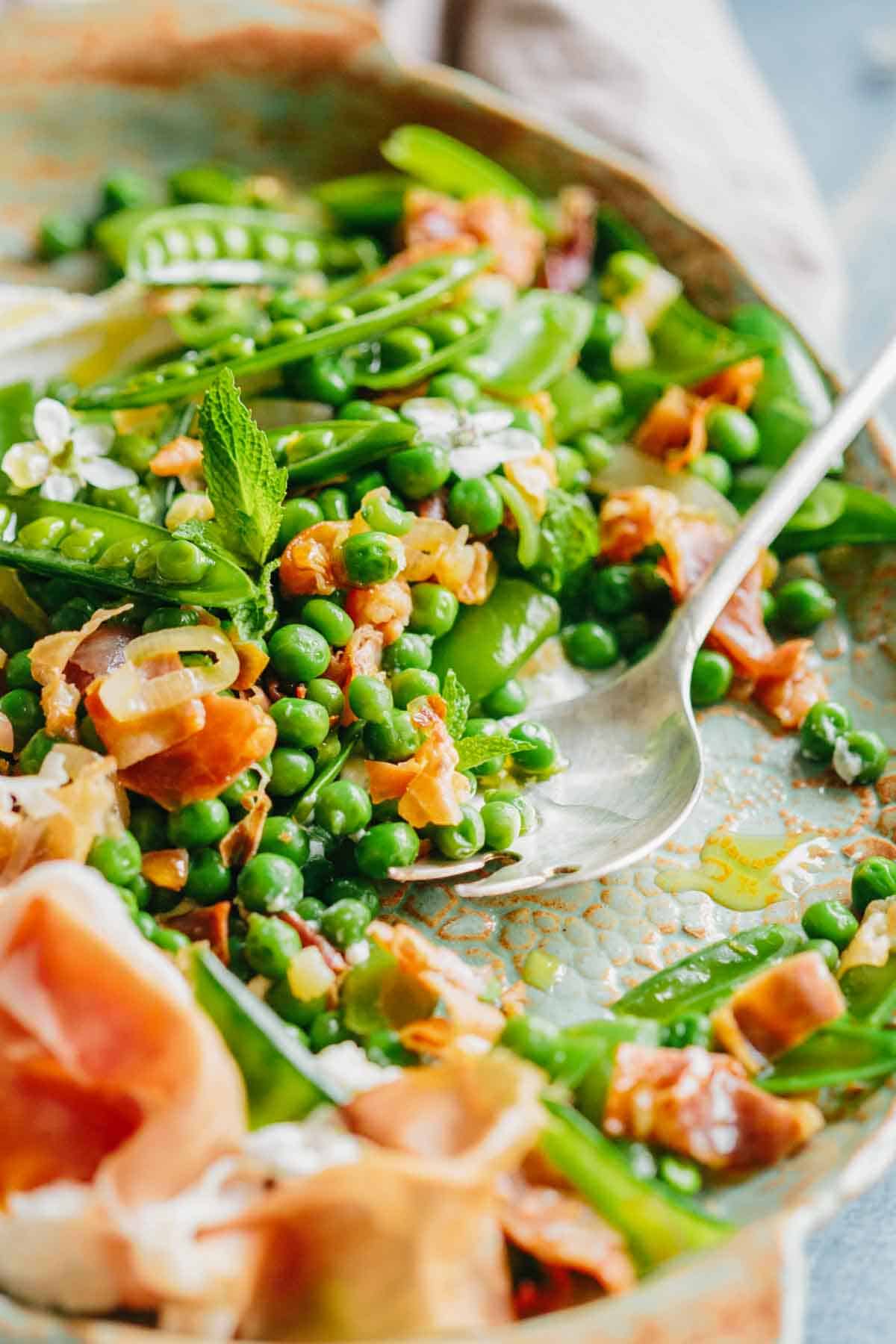 Close-up of a spring pea dish in a copper pan showing vibrant green peas, snow peas, crispy prosciutto bits, and fresh mint leaves with a serving spoon.