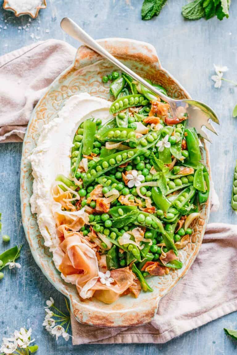 Overhead view of an elegant oval serving platter with bright green peas and snap peas topped with prosciutto and fresh herbs, served with a swirl of creamy ricotta cheese.