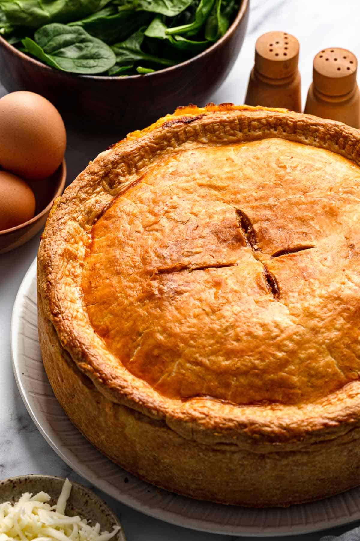 Golden-brown deep dish pie with tall fluted edges on a gray serving plate with a bowl of fresh spinach, eggs, and grated cheese visible in the background.