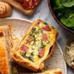 Overhead view of a white plate with slices of Italian Easter pie, surrounded by sliced salami, fresh spinach and shredded cheese on a marble surface.