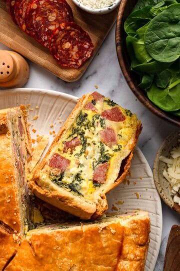 Overhead view of a white plate with slices of Italian Easter pie, surrounded by sliced salami, fresh spinach and shredded cheese on a marble surface.