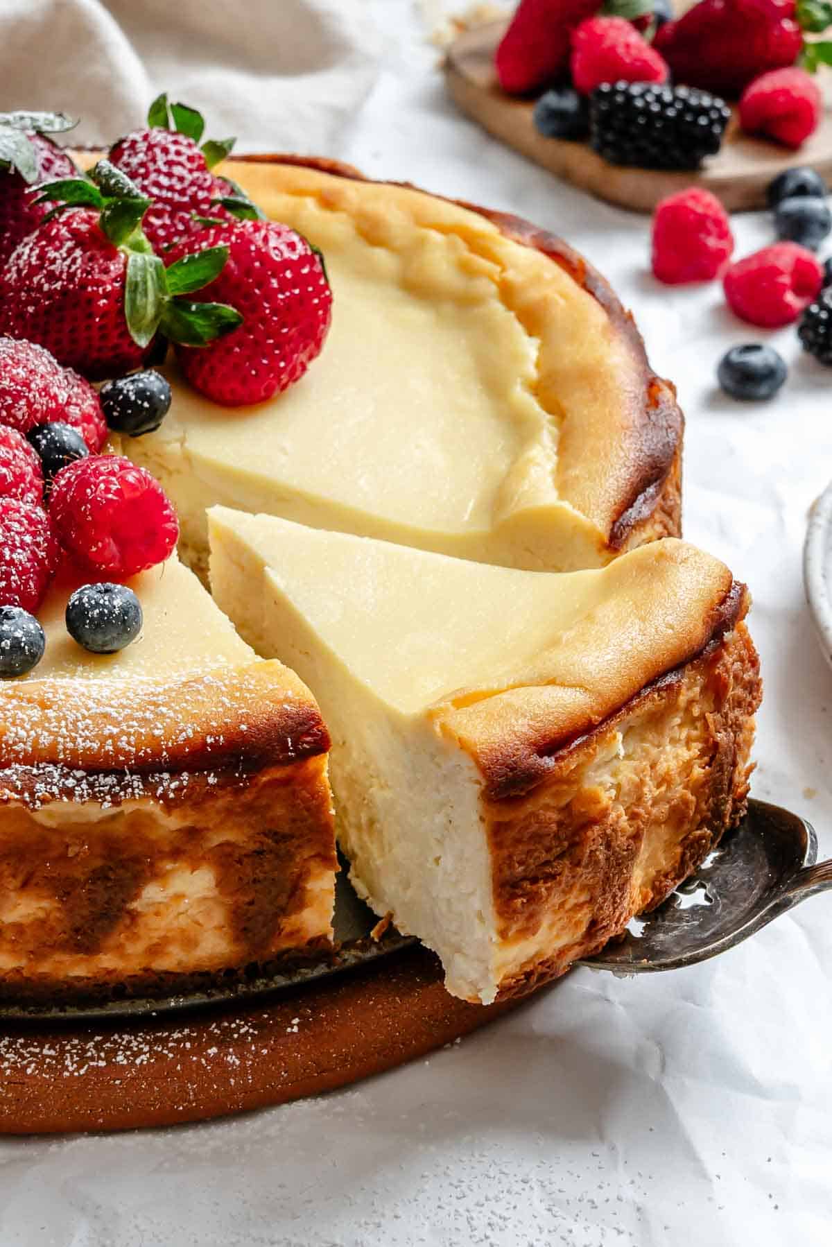 Close-up view of a ricotta cheesecake with a slice being removed, showing the smooth, dense texture. The cake is decorated with fresh berries and the golden brown crust is visible along the sides.