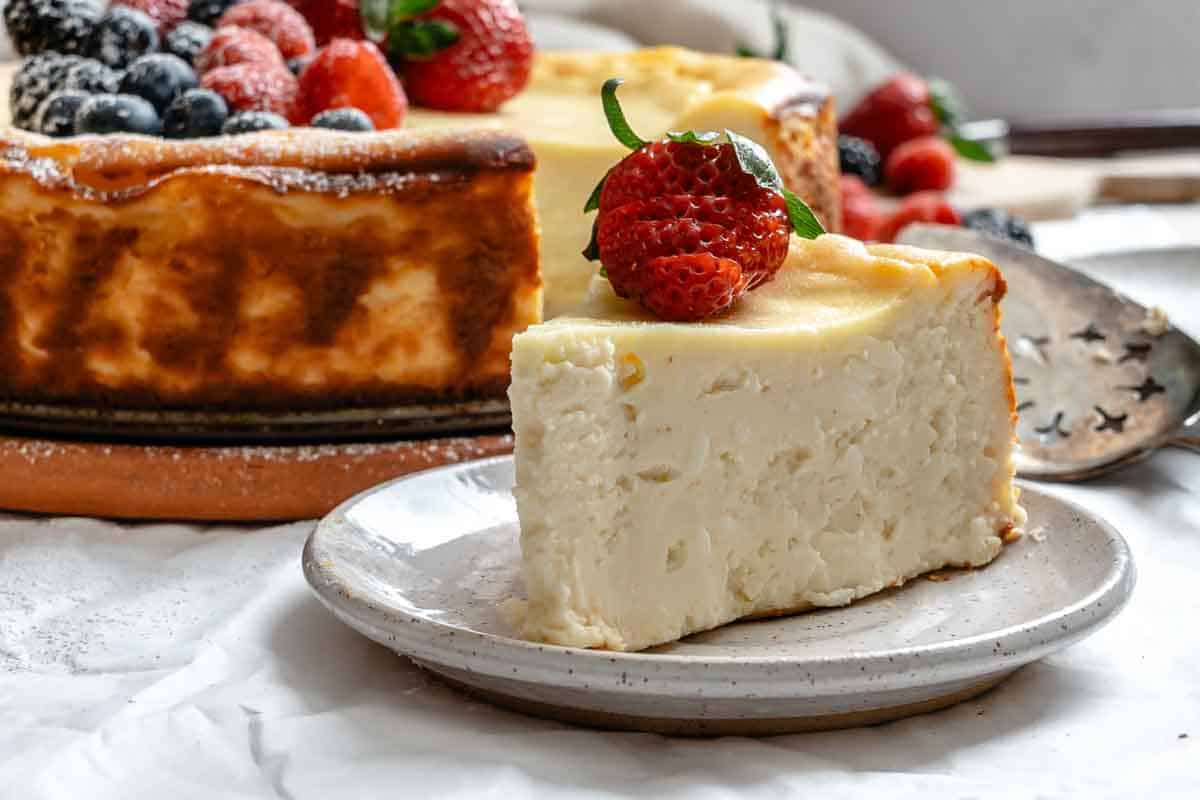 Close-up of a slice of ricotta cheesecake on a speckled white plate with a fresh strawberry on top. The whole cheesecake topped with mixed berries is visible in the background.