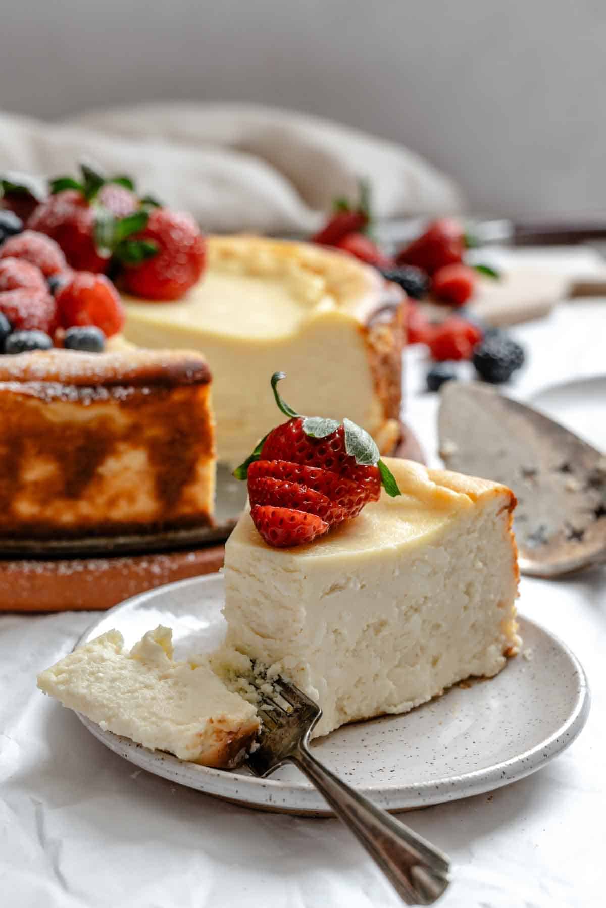 Close-up of a slice of ricotta cheesecake on a white plate with a bite taken out on a fork. A fresh strawberry garnishes the slice, and the rest of the berry-topped cheesecake is visible in the soft-focus background.
