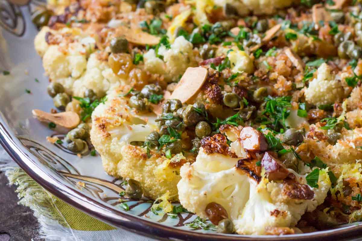 Close-up view of roasted cauliflower with crunchy breadcrumb topping, capers, and sliced almonds on a decorative plate with a patterned rim.