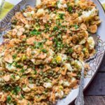 Overhead view of a complete dish of roasted cauliflower with breadcrumbs, capers, herbs and almonds served on a decorative platter with a silver serving spoon.