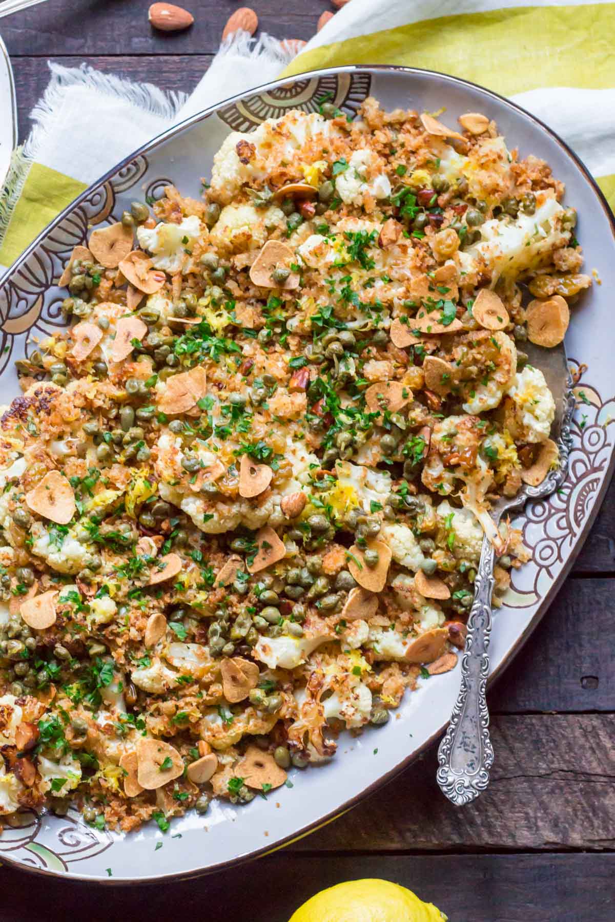 Overhead view of a complete dish of roasted cauliflower with breadcrumbs, capers, herbs and almonds served on a decorative platter with a silver serving spoon.