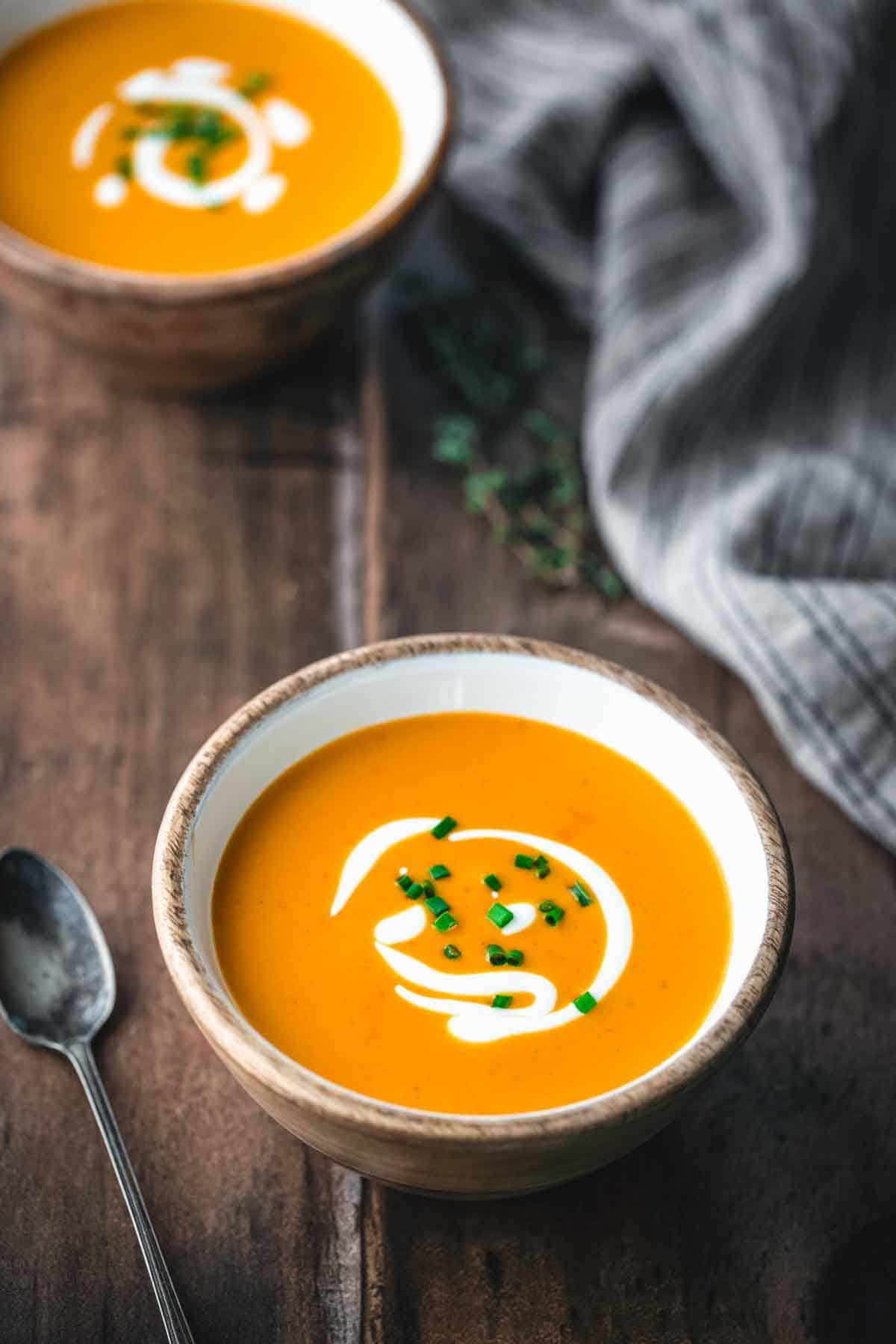 Close-up of creamy orange shrimp bisque in a wooden-rimmed bowl garnished with a decorative swirl of cream and minced chives. A spoon is visible alongside with a gray linen napkin in the background.
