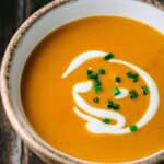 Extreme close-up of shrimp bisque in a wooden-rimmed white bowl showing the smooth, velvety orange-colored soup with decorative cream swirl and bright green chive garnish against a dark wooden background.