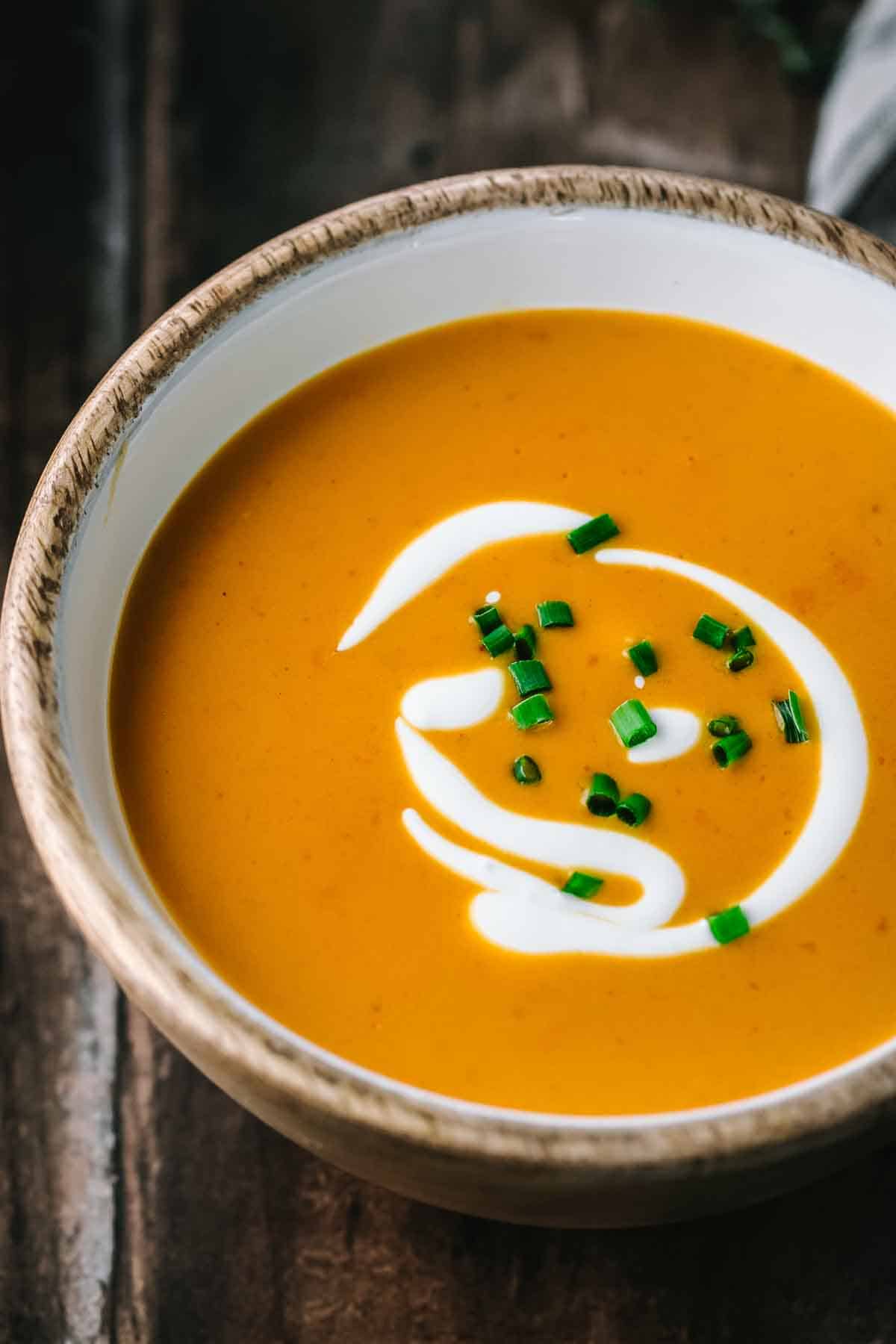 Extreme close-up of shrimp bisque in a wooden-rimmed white bowl showing the smooth, velvety orange-colored soup with decorative cream swirl and bright green chive garnish against a dark wooden background.