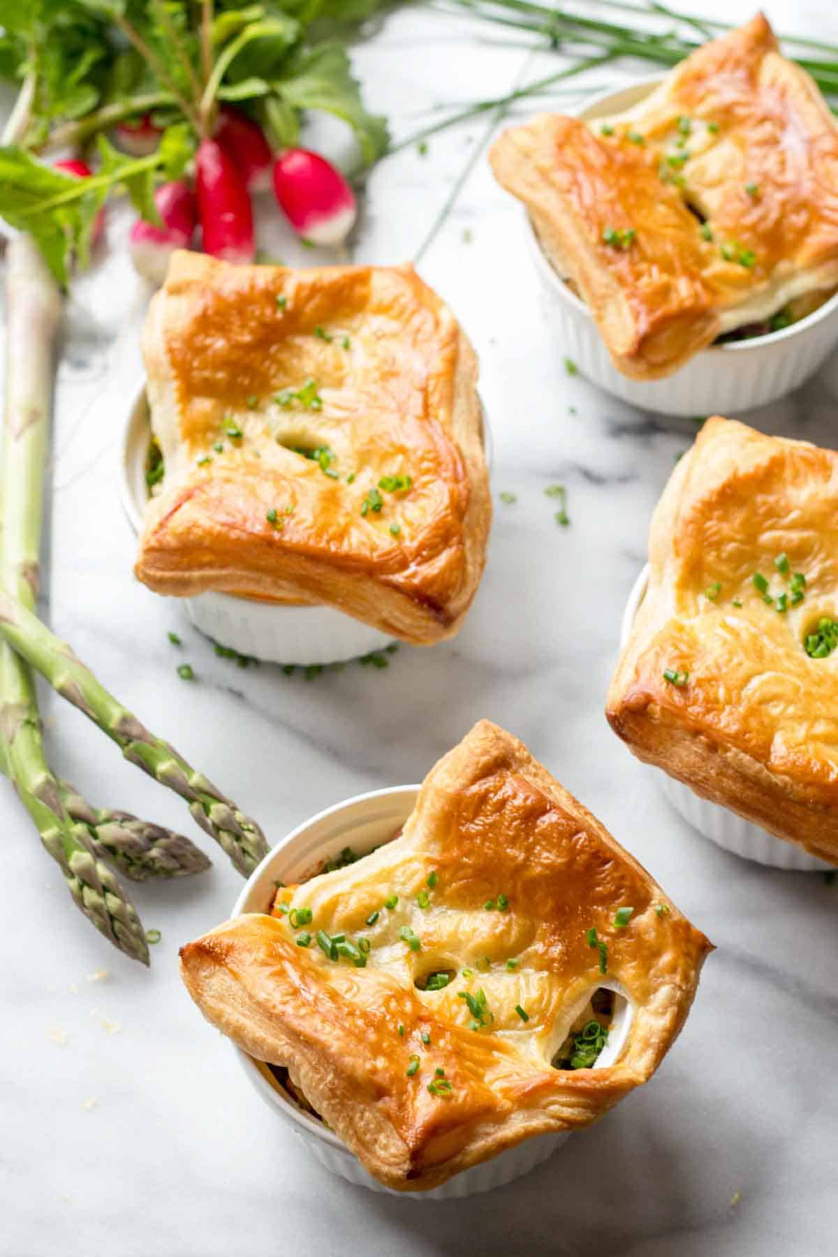 Four golden-brown chicken pot pies in white ramekins on a marble surface, garnished with fresh chives. Fresh asparagus spears and radishes are visible as decorative elements. The puff pastry tops are beautifully puffed and flaky.