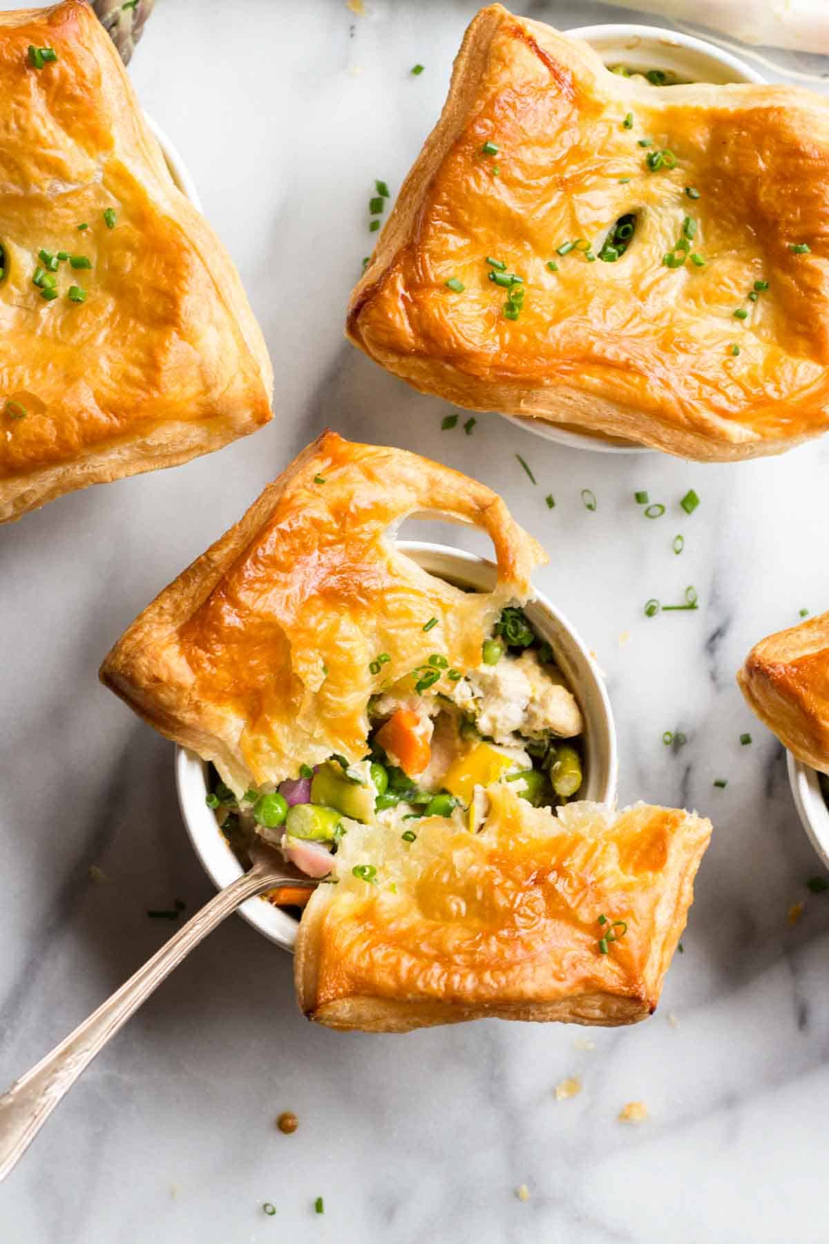Overhead view of an individual chicken pot pie with part of the golden pastry top broken and a spoonful of filling being lifted out. The filling shows pieces of chicken, colorful vegetables, and a creamy sauce.