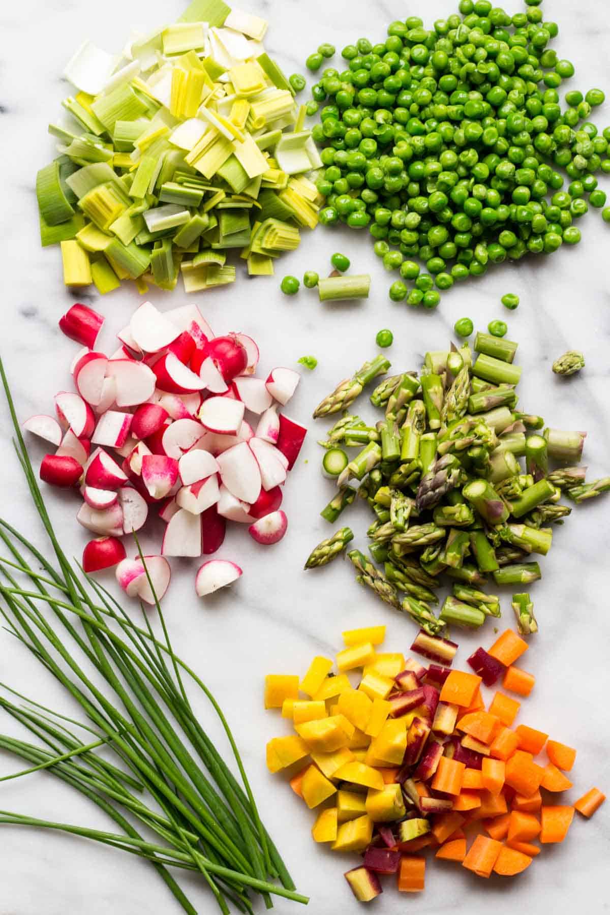 Ingredients for spring chicken pot pie arranged on white marble: chopped leeks, green peas, radishes, asparagus pieces, diced colorful bell peppers and carrots, and fresh chives. The vegetables are arranged in separate piles showcasing the vibrant spring colors.