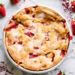 Top-down view of a whole strawberry pie in a white baking dish. The golden crust has a dusting of sugar and shows bubbling red strawberry filling. Fresh strawberries and delicate pink blossoms surround the pie on a marble surface.