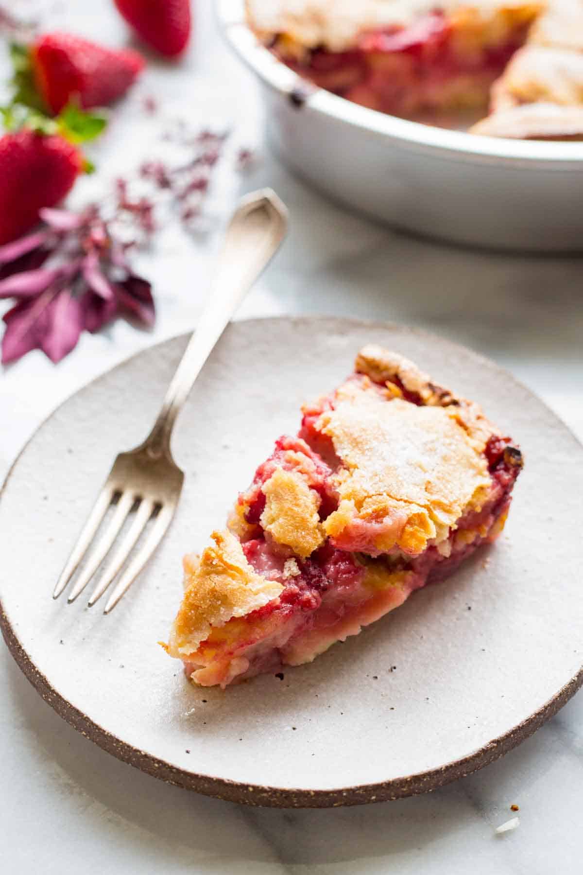 Close-up of a strawberry pie slice on a white plate with a silver fork. The pie has a flaky golden crust and bright red strawberry filling. Fresh whole strawberries and purple flowers are arranged around the plate on a marble surface.