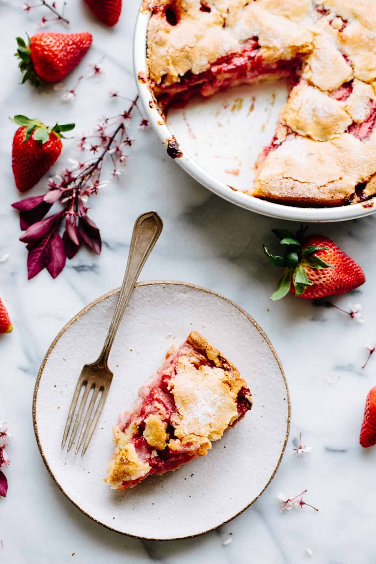Overhead view of a partially eaten strawberry pie in a white pie dish with a slice removed and served on a white plate. The scene is decorated with fresh strawberries and pink spring blossoms on a marble countertop.