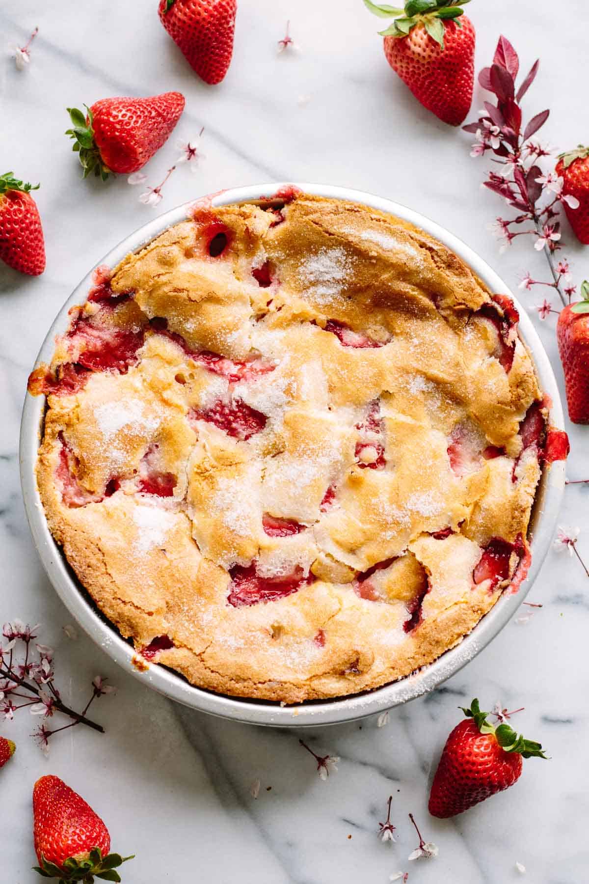 Top-down view of a whole strawberry pie in a white baking dish. The golden crust has a dusting of sugar and shows bubbling red strawberry filling. Fresh strawberries and delicate pink blossoms surround the pie on a marble surface.