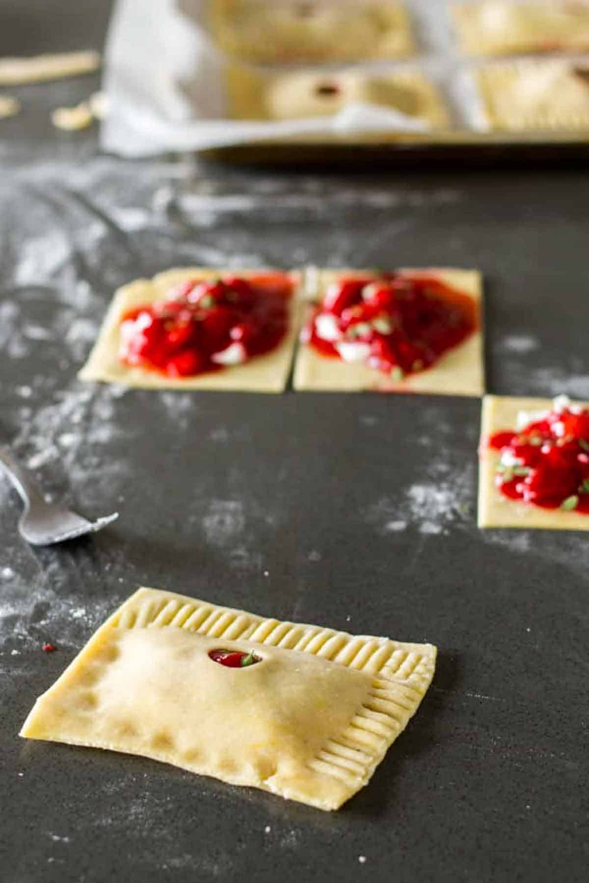 Squares of pastry dough on a dark surface. Some squares have red strawberry filling on top, while one completed pastry in the foreground has been sealed with crimped edges and has a small vent hole.