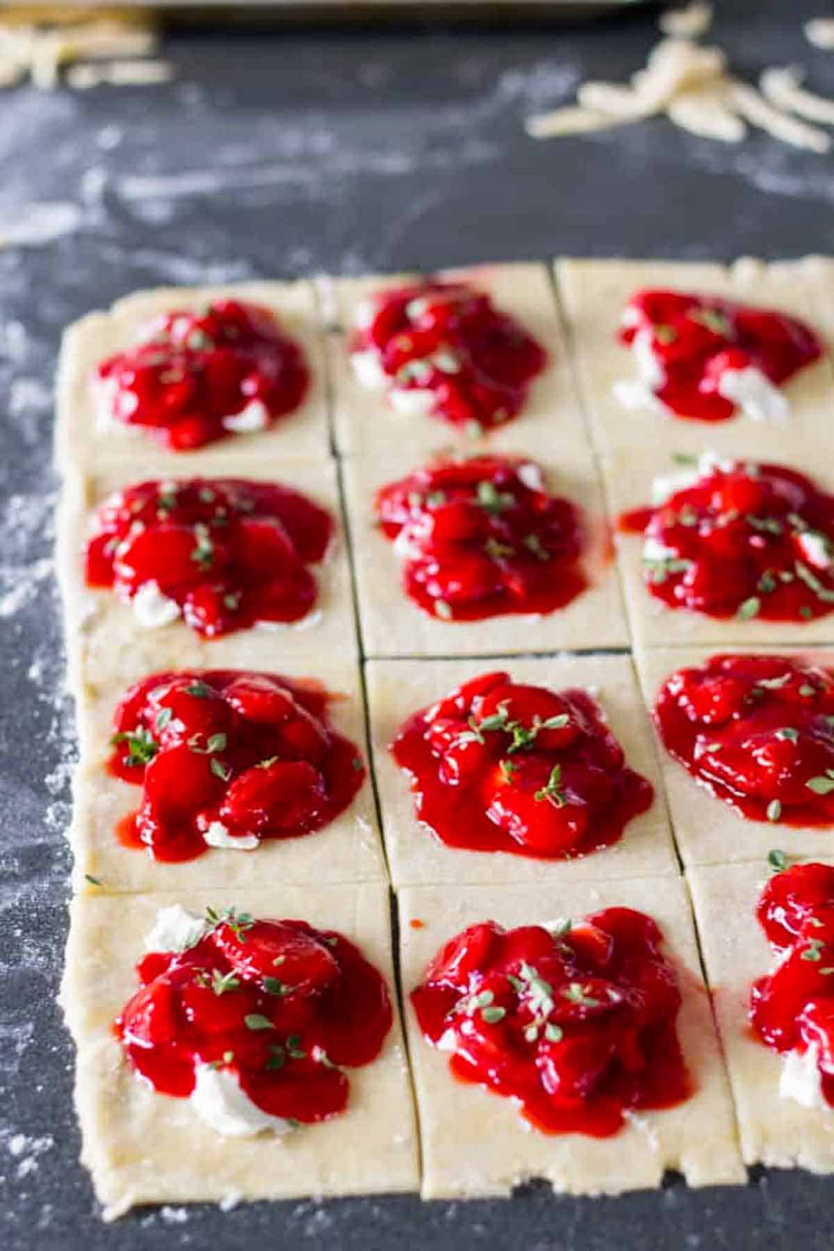 A grid of pastry squares, each topped with a dollop of bright red strawberry filling and what appears to be herbs or green garnish on a floured dark surface.
