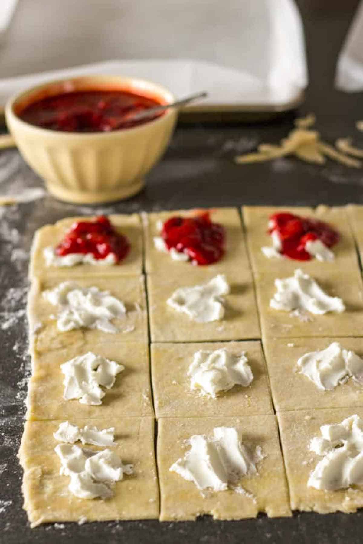 Captures the assembly process with squares of dough laid out on a dark surface. The bottom squares have white goat cheese dollops, while the top row has both cheese and strawberry filling. A small yellow bowl of strawberry filling is visible in the background.