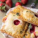 Several finished hand pies on a decorative plate with fresh strawberries in the background. The pastries are golden brown with vent holes showing the red filling inside, and are garnished with small herbs.