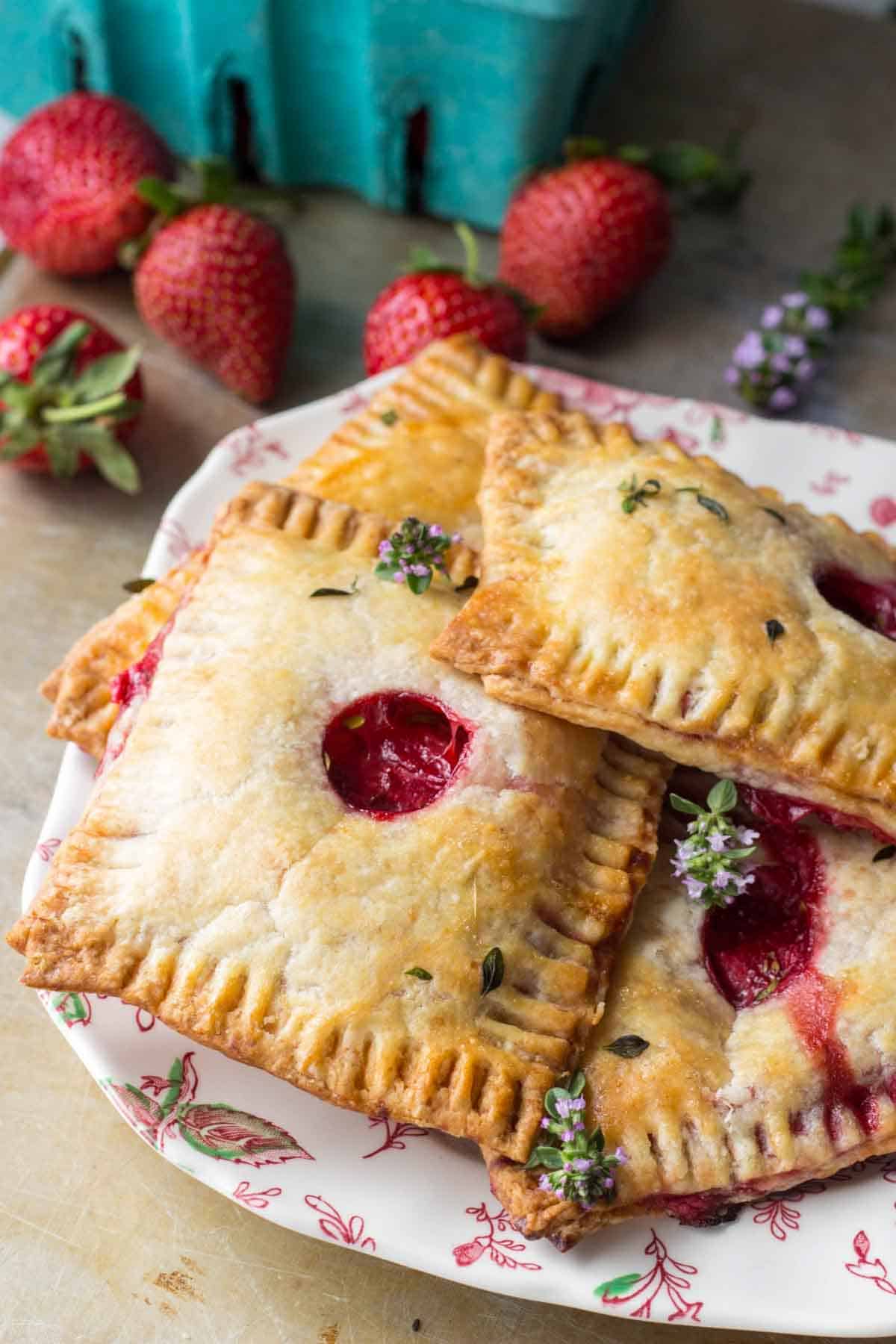 Several finished hand pies on a decorative plate with fresh strawberries in the background. The pastries are golden brown with vent holes showing the red filling inside, and are garnished with small herbs.
