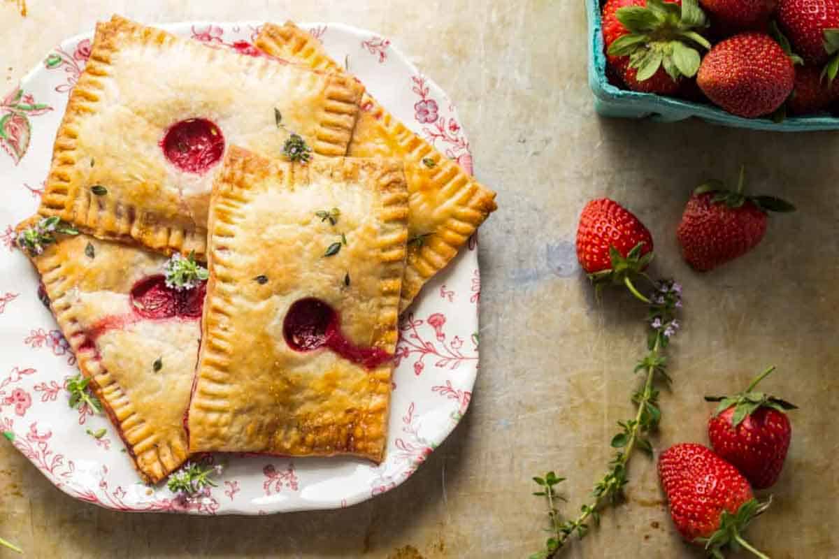 Presentation shot showing the finished pastries on a plate with a decorative floral rim. Fresh strawberries are arranged around the plate and in a teal container visible at the edge of the frame.