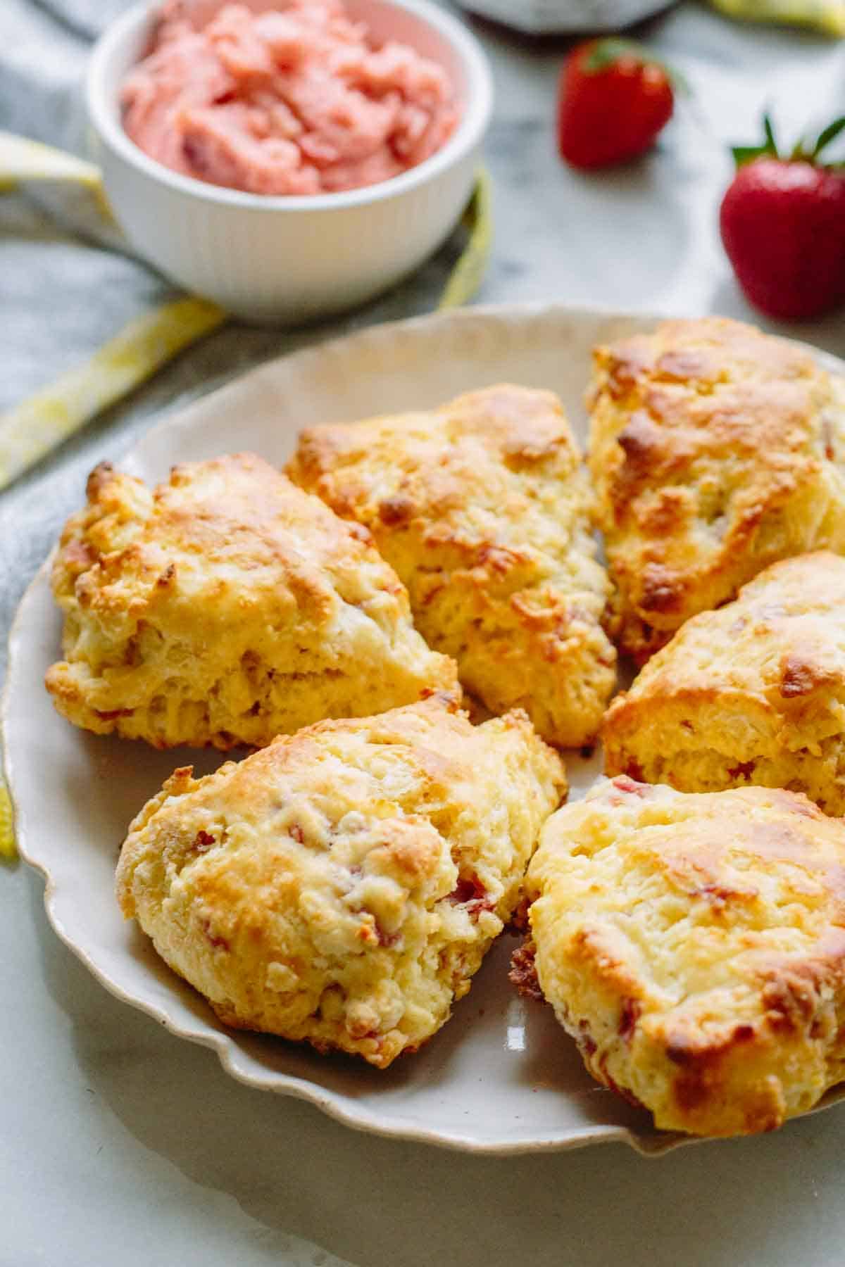 Golden-brown goat cheese and prosciutto scones arranged on a white scalloped plate. A small bowl of pink strawberry butter is visible in the background along with fresh strawberries on a marble countertop.