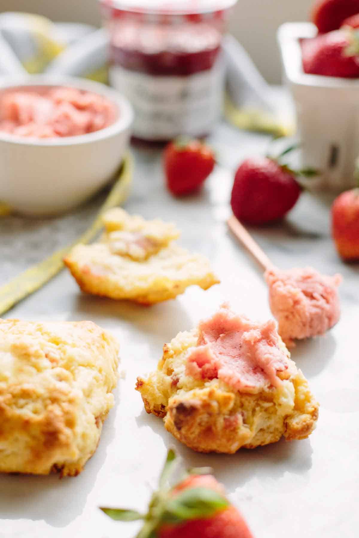 Close-up of goat cheese prosciutto scones on a white surface with strawberry butter spread on one scone. Fresh strawberries, a jar of preserves, and a bowl of strawberry butter are visible in the soft-focus background.
