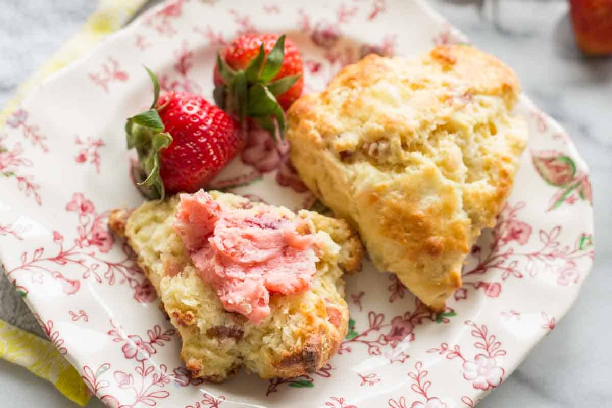 Two goat cheese prosciutto scones on a decorative plate with pink floral pattern. One scone is topped with strawberry butter, and fresh strawberries garnish the plate. The scones show a golden crust with a tender interior.