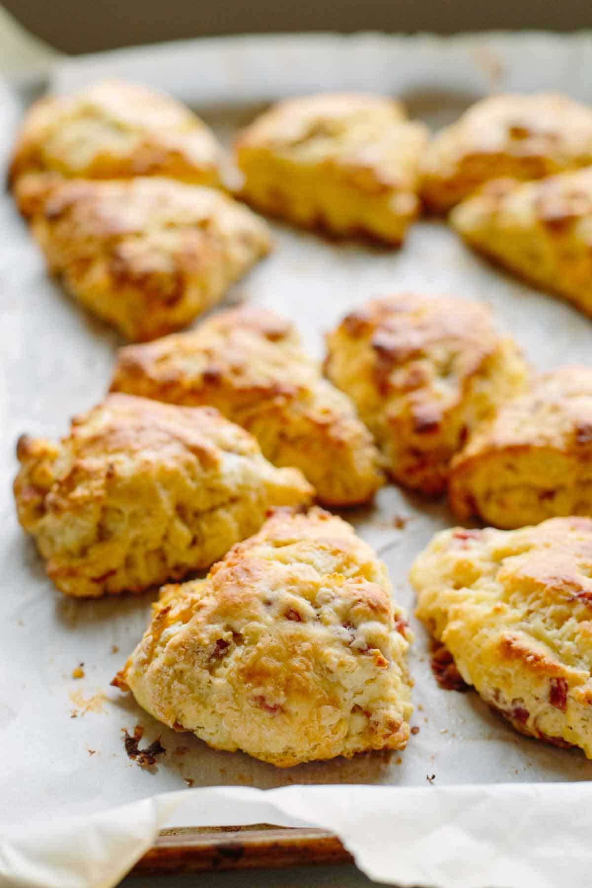 Freshly baked goat cheese and prosciutto scones on a parchment-lined baking sheet. The scones have a rustic, golden appearance with visible bits of prosciutto throughout and slightly crisp, browned edges.