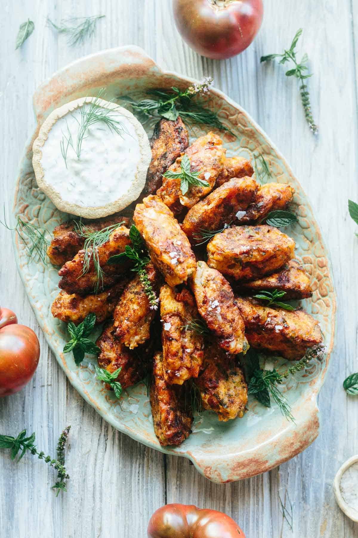 Golden tomato fritters are artfully arranged on a ceramic platter, garnished with fresh herbs and served alongside a rustic bowl of creamy tzatziki. The light wood backdrop and scattered heirloom tomatoes evoke a summery, garden-fresh feel.