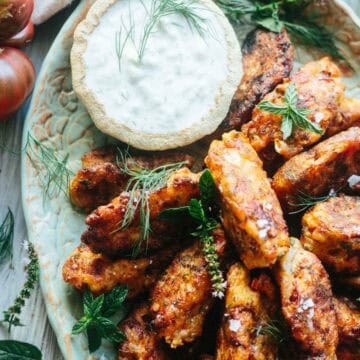 A close-up of crispy tomato fritters stacked generously around a dish of herbed tzatziki sauce. Flecks of sea salt and sprigs of dill and mint highlight the dish’s vibrant, savory charm.