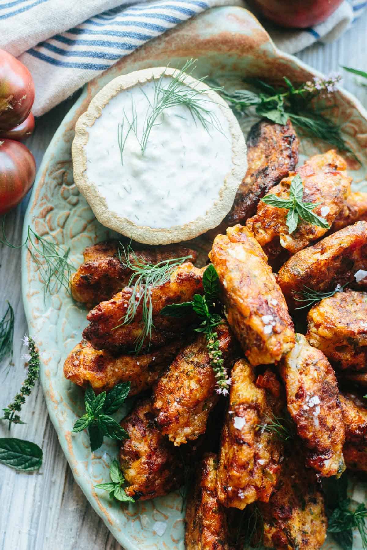 A close-up of crispy tomato fritters stacked generously around a dish of herbed tzatziki sauce. Flecks of sea salt and sprigs of dill and mint highlight the dish’s vibrant, savory charm.