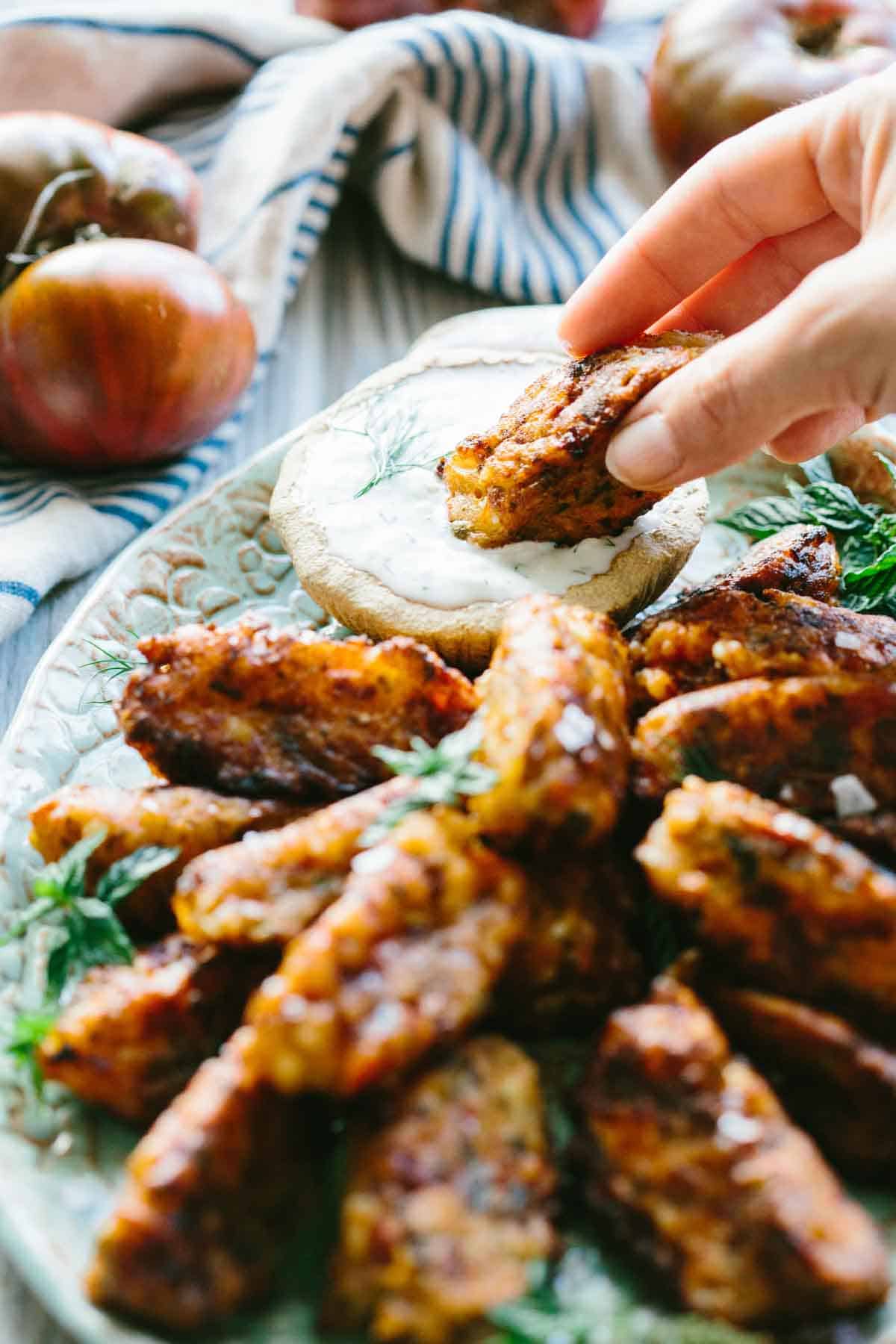 A hand reaches in to dip a hot, crispy tomato fritter into a cool dollop of tzatziki. The plate overflows with more golden fritters, hinting at irresistible texture and flavor in every bite.