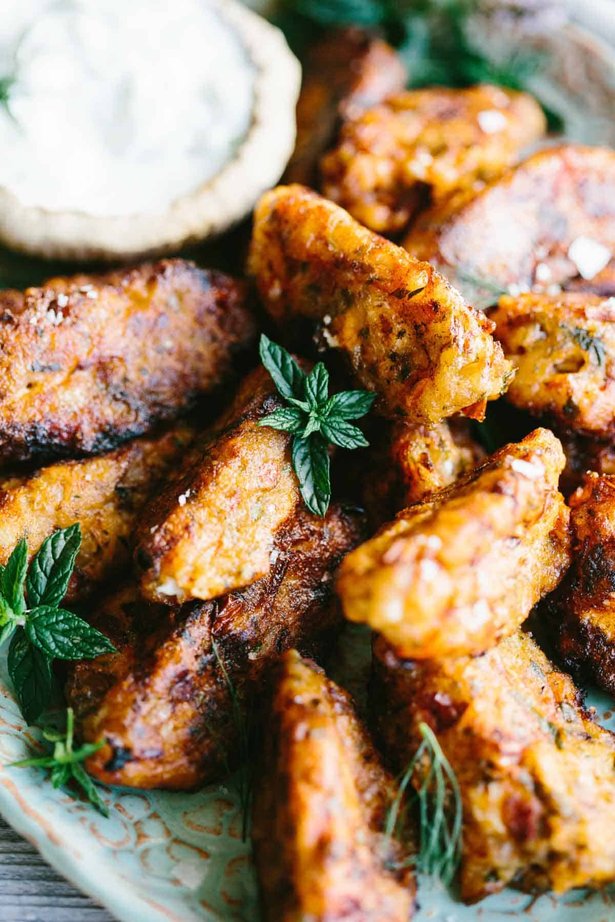 Tomato fritters are stacked tightly in a rustic ceramic dish, their crisp edges and golden color glistening beside a bowl of tzatziki. A sprig of mint adds a pop of freshness to the composition.