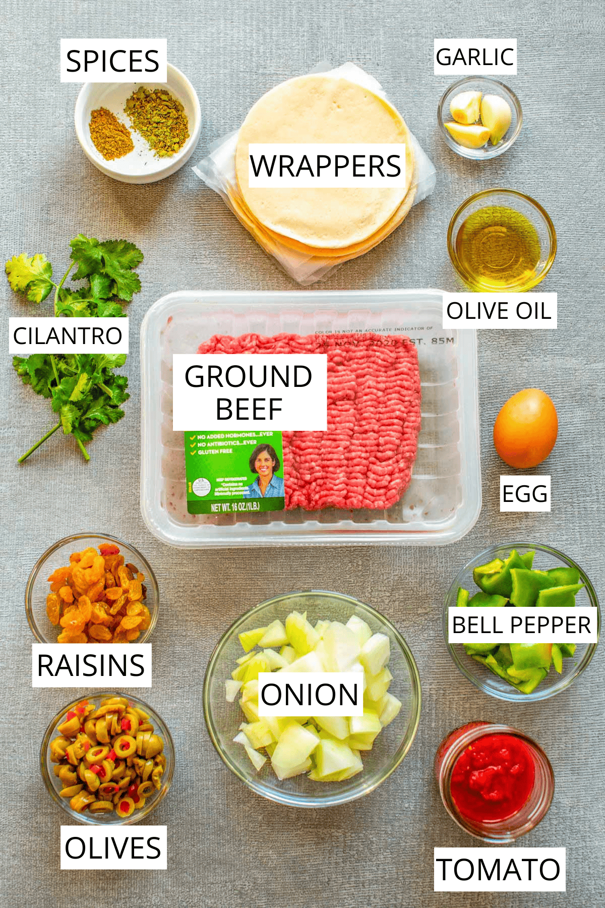 Overhead view of ingredients for making picadillo empanadas, including: packaged ground beef, pre-made empanada discs, spice mixtures, chopped green peppers, diced onions, sliced olives, golden raisins, tomato sauce, olive oil, garlic, and fresh cilantro. Each component is neatly arranged in separate bowls on a gray textured surface.
