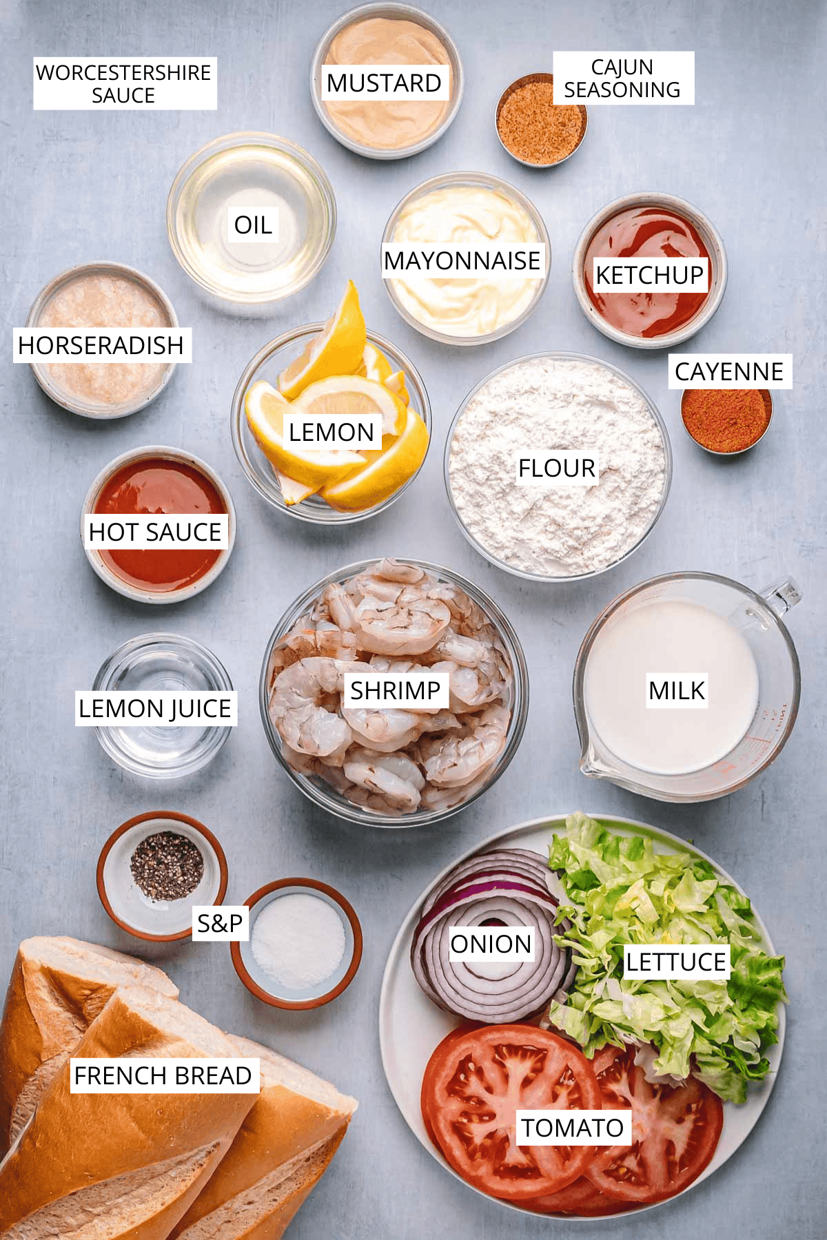 Flat lay of labeled ingredients for a shrimp po’boy, including raw shrimp, French bread, lettuce, tomato, red onion, milk, flour, and condiments like mayonnaise, ketchup, mustard, hot sauce, and seasonings.