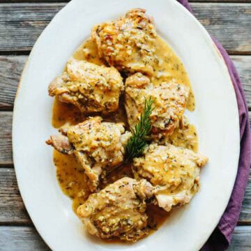 Overhead view of an oval white serving platter containing chicken thighs coated in a golden sauce with visible herb specks. A sprig of rosemary sits in the center, and the dish is presented on a rustic wooden surface with a purple napkin alongside.