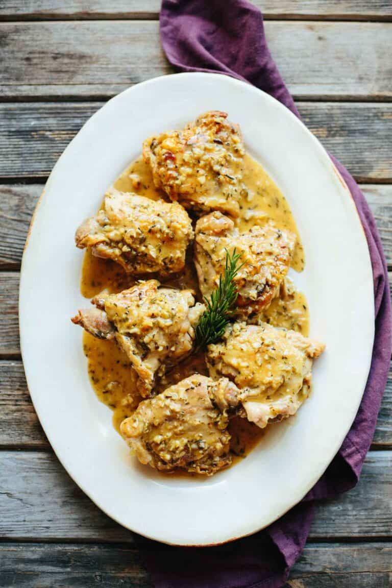 Overhead view of an oval white serving platter containing chicken thighs coated in a golden sauce with visible herb specks. A sprig of rosemary sits in the center, and the dish is presented on a rustic wooden surface with a purple napkin alongside.