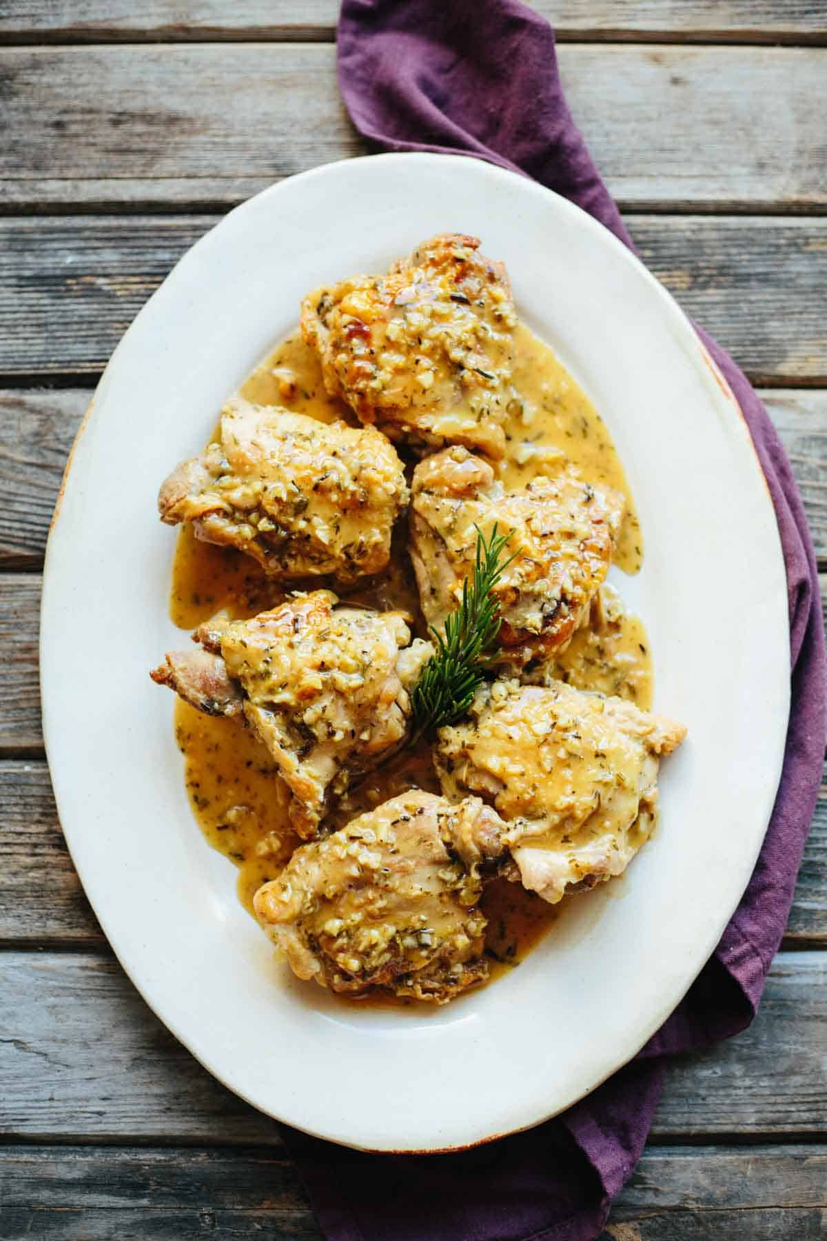 Overhead view of an oval white serving platter containing chicken thighs coated in a golden sauce with visible herb specks. A sprig of rosemary sits in the center, and the dish is presented on a rustic wooden surface with a purple napkin alongside.