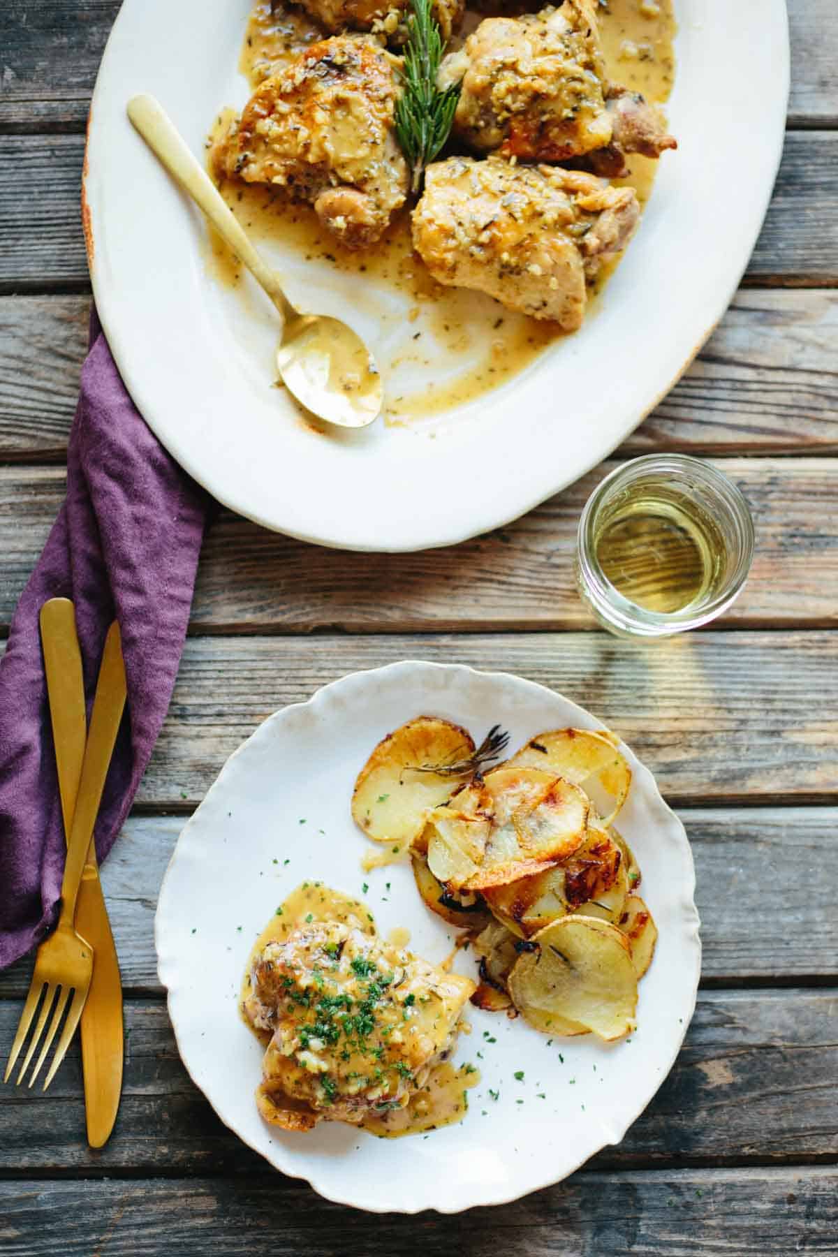 Table setting showing two plates - the main platter of chicken cacciatore with rosemary garnish above, and a single serving with roasted potatoes below. A glass of white wine and gold utensils complete the elegant presentation on a wooden table.