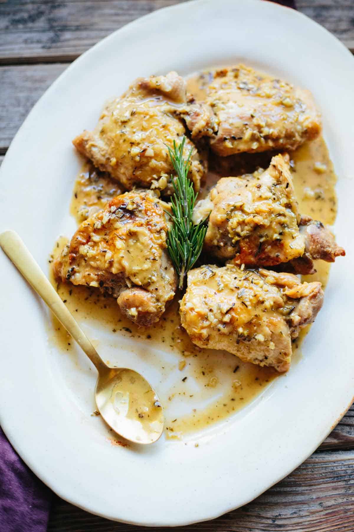 Focused overhead view of chicken thighs arranged on a white oval platter, swimming in a light-colored, herb-infused sauce. A gold spoon rests alongside, ready for serving, with the rosemary sprig providing a decorative touch.