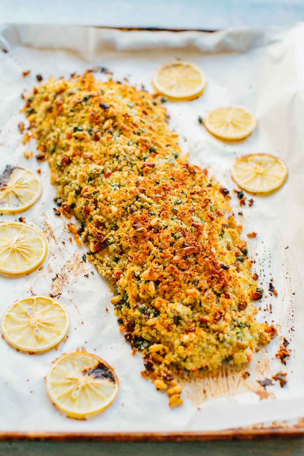 A salmon fillet on parchment paper, coated with golden-brown breadcrumb topping flecked with herbs, nuts, and raisins. Thin lemon slices arranged around the fish add brightness to the presentation.