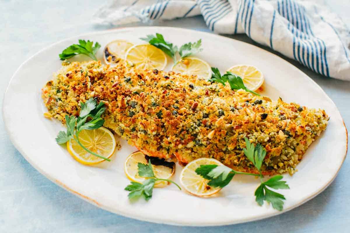 Beautifully plated breadcrumb-crusted salmon on a white serving dish, garnished with charred lemon slices and fresh parsley sprigs. The golden topping displays a textured blend of Sicilian-inspired ingredients.